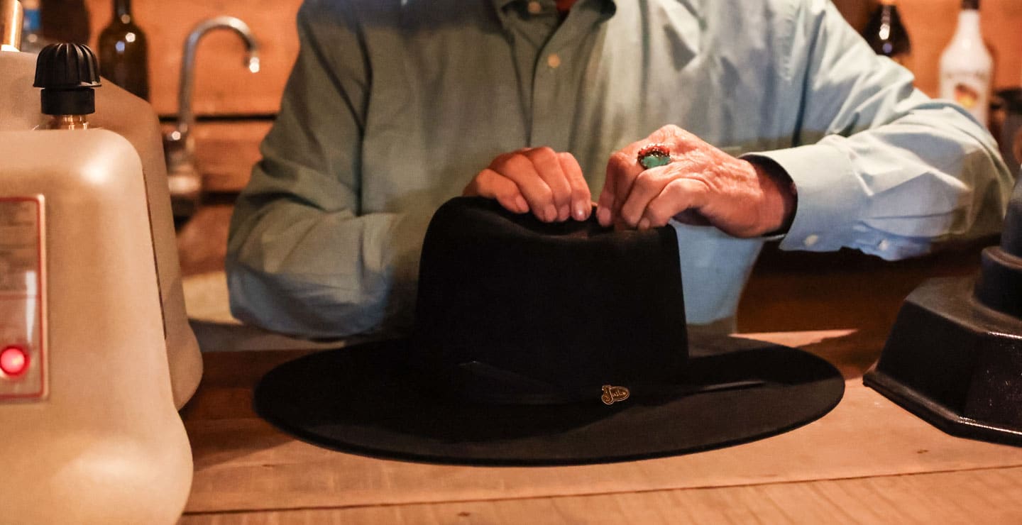 A man with his hands on a black hat on the counter, shaping a hat.
